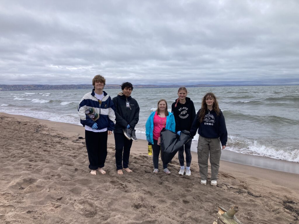 Five people standing on a sandy beach with a lake and cloudy sky in the background. They are wearing jackets and holding items, possibly for a beach cleanup activity. The group appears to be friends, and they are smiling while facing the camera.