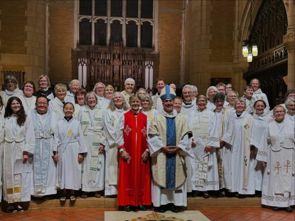 A group of clergy members stand together in a church. They are dressed in a variety of liturgical garments, primarily white robes; one individual wears a red robe, and another is adorned in a blue and white outfit with a mitre. They pose for a group photo, all smiling.