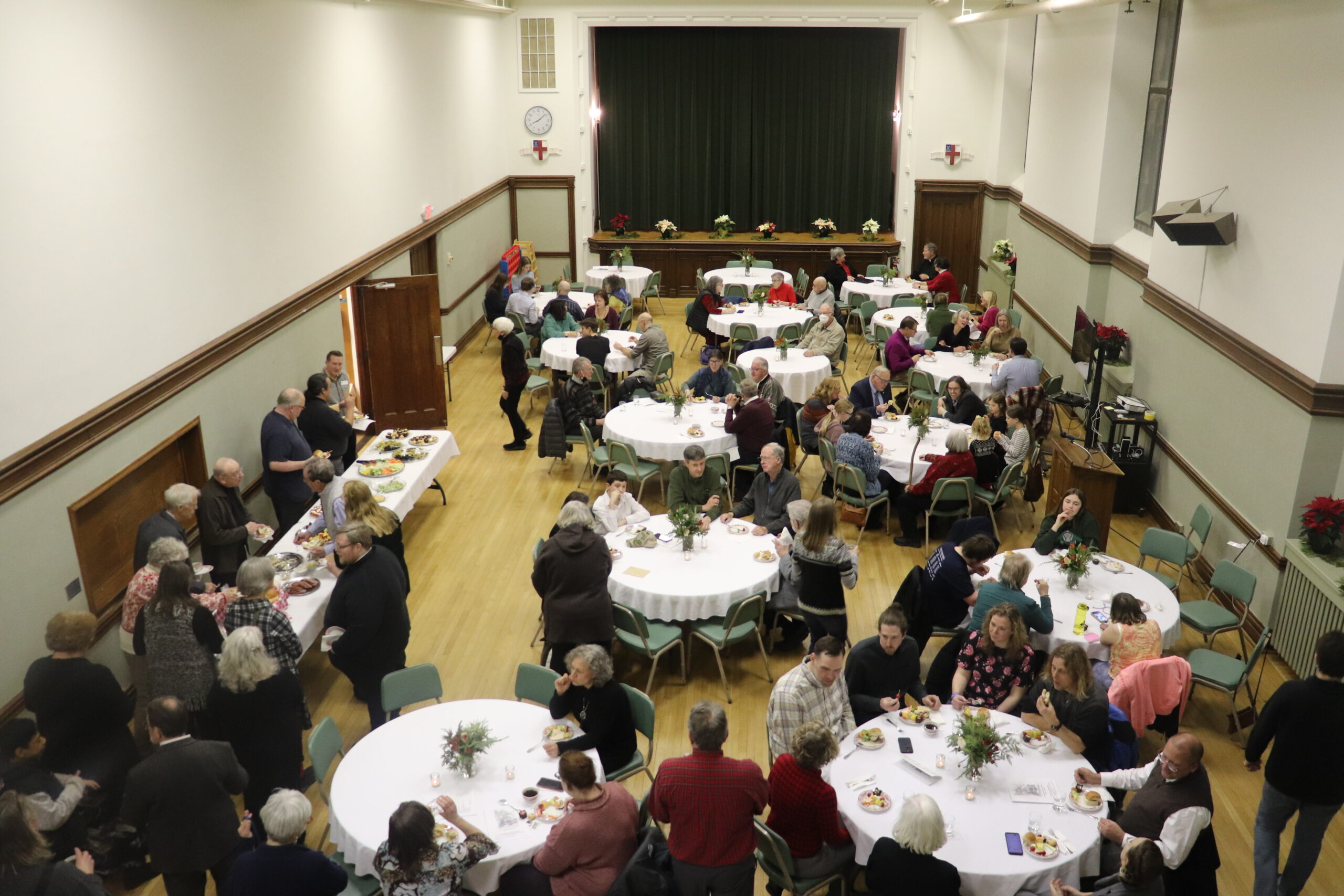 A large community hall hosts a gathering with several round tables and numerous guests seated and standing. People are engaged in conversations and enjoying food. The room is decorated with festive flowers and the atmosphere appears lively and social.